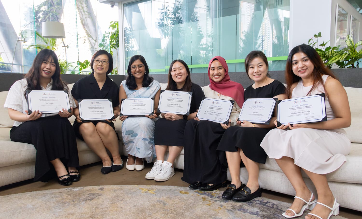 Seven women seated on a sofa, smiling and holding framed certificates in a bright office with plants.